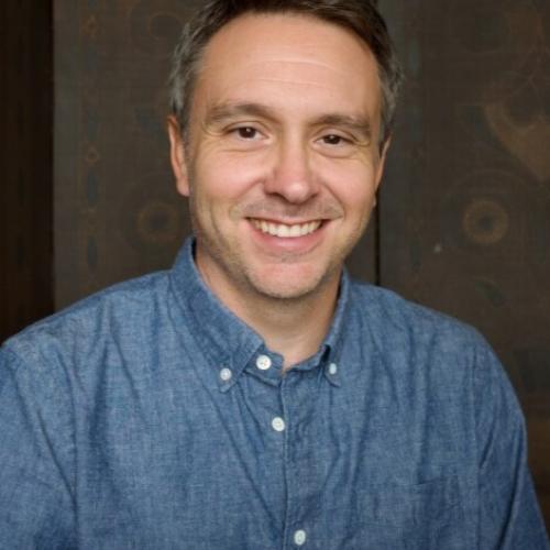 Headshot of C.J. Alvarez. He wears a denim blue button up shirt and is pictured behind a brownish background. 